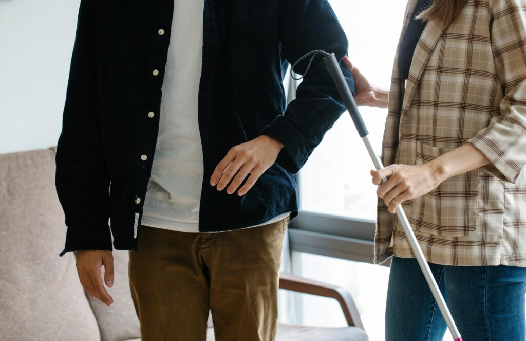 Two adults providing support indoors with a walking stick, displaying teamwork and care.